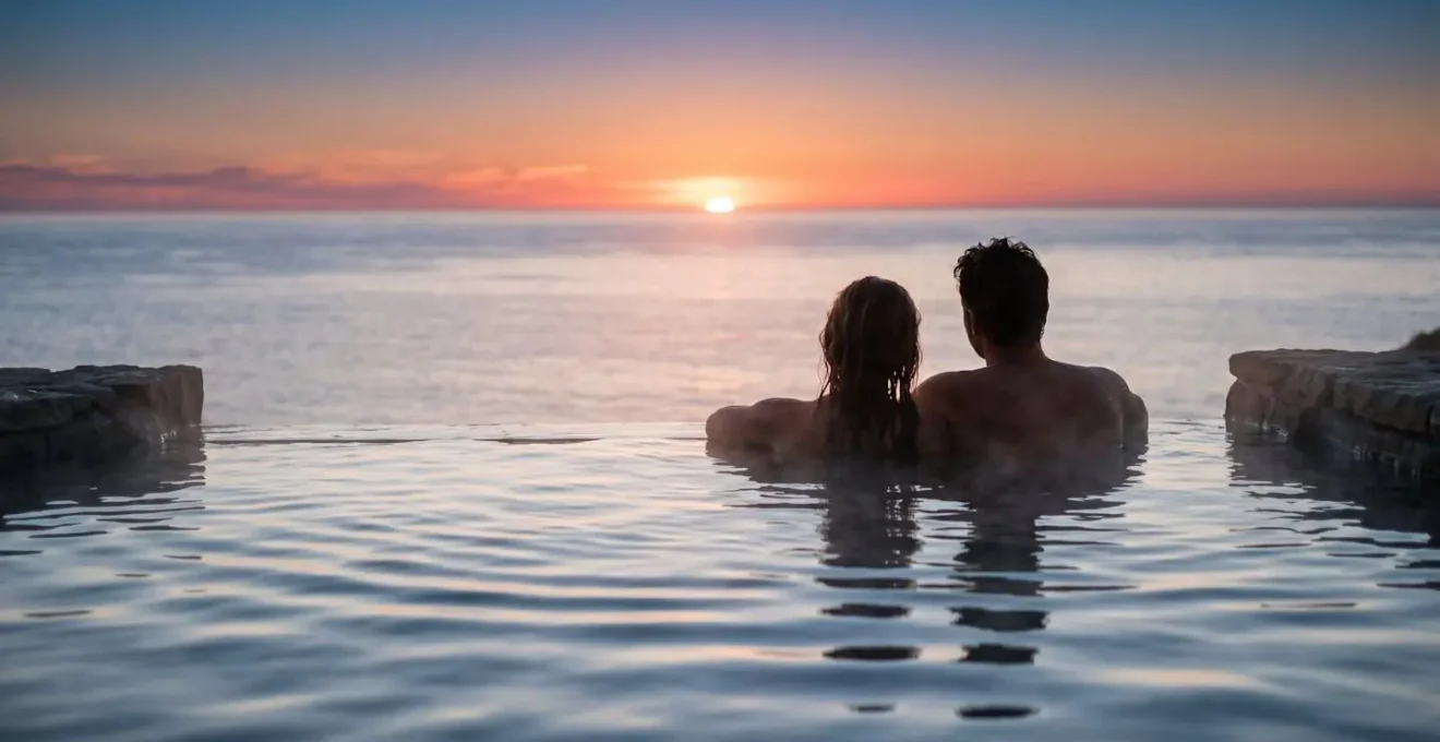 Couple admirant un coucher de soleil depuis une piscine à débordement face à l'océan, évoquant l'expérience du Sky Lagoon.